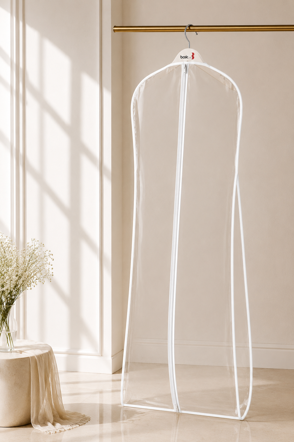A clear plastic garment storage bag with a white zipper and trim, hanging from a gold hanger on a clothing rack in a sunlit beige room.